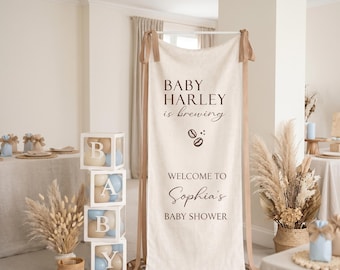 May include: A tall, rectangular, off-white fabric banner hanging from a white frame with tan ribbons. The text reads: BABY HARLEY is brewing, two coffee bean icons, WELCOME TO, Sophia's, BABY SHOWER. Next to it are four stacked clear boxes spelling BABY.