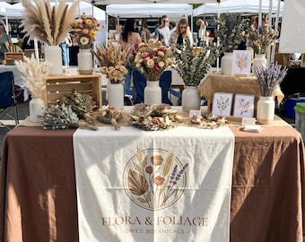 May include: A market stall display featuring various dried flower arrangements in white ceramic vases and bundles of dried botanicals. A cream-colored table runner displays the text Flora & Foliage Dried Botanicals with a circular floral graphic.