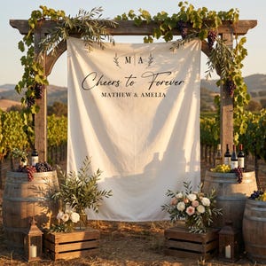 May include: A large off-white fabric backdrop hanging from a wooden archway, featuring the text "Cheers to Forever" and "MATHEW & AMELIA" with "M A" initials. The display includes two wooden wine barrels, crates, floral arrangements, and grape vines.