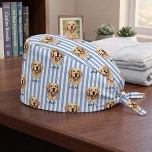 May include: A scrub cap featuring a light blue and white vertical striped pattern with repeating illustrations of golden retriever dog faces and the word Lucky printed in black text below each dog face. The cap includes fabric ties on the side.