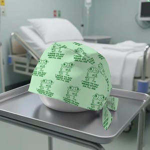 May include: A light green surgical scrub cap with a repeating pattern of cartoon frogs and the text "I HAVE TO SAY WEIRD STUFF OR I'LL DIE." The cap is tied at the back and sits on a white bowl.