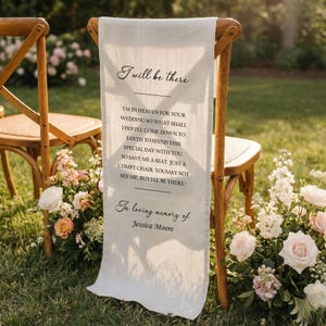 May include: A cream-colored fabric chair sash with the text "I will be there" and a poem about a wedding. The sash is draped over a wooden chair, with floral arrangements and greenery in the background.