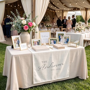May include: A beige wedding welcome table featuring a runner printed with "The Anderson EST. 2026". The table holds a large vase with eucalyptus and pink roses, several framed photographs, a guest book, a gold glass box, and small candles.