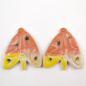 Puede incluir: Dos adornos de cerámica con forma de polilla con un esmalte amarillo, naranja y blanco. Los adornos tienen una superficie texturizada y detalles negros.