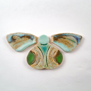 May include: A ceramic art piece with a butterfly-like design. The piece features a central body with two green circles and two wing-shaped elements. The colors include shades of blue, brown, and green.