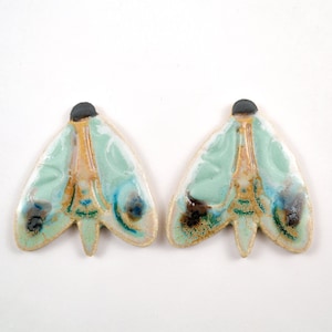 Op de afbeelding: Een paar keramische oorringen in de vorm van een mot met een lichtgroene, blauwe en bruine glazuur. De oorringen hebben een klein zwart accent aan de bovenkant.