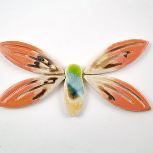 Puede incluir: Una mariposa de cerámica con alas de color naranja, blanco y marrón. El cuerpo de la mariposa es de color azul claro y verde.