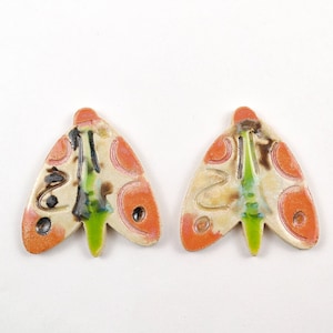 Puede incluir: Dos adornos de cerámica con forma de mariposa con base blanca, detalles verdes, naranjas y marrones.