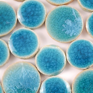 Puede incluir: Una colección de azulejos redondos de cerámica azul verdoso con un acabado de esmalte craquelado. Los azulejos tienen un borde beige claro.