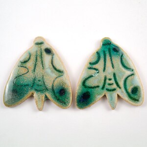 Puede incluir: Dos adornos de cerámica con forma de mariposa con un esmalte verde y blanco. Las mariposas tienen un diseño detallado con un fondo blanco y líneas y remolinos verdes.