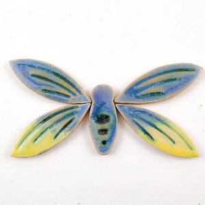 Può includere: Una piastrella di mosaico in ceramica a forma di farfalla con accenti blu, gialli e verdi. La farfalla ha un corpo segmentato e ali con un design dettagliato.