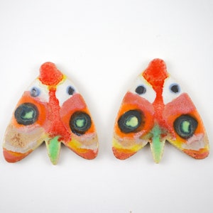 Puede incluir: Dos pendientes de cerámica con forma de mariposa con detalles coloridos, incluyendo rojo, naranja, amarillo, verde, azul y blanco. Las mariposas tienen una superficie texturizada y tienen forma de polilla.