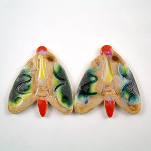 Puede incluir: Dos adornos de cerámica con forma de mariposa con base blanca y esmalte colorido. El esmalte presenta detalles en verde, azul, amarillo y rojo. Los adornos miden aproximadamente 5 cm de largo.