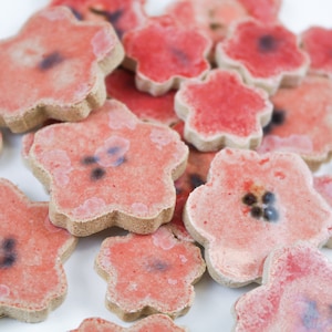 May include: A cluster of ceramic flower-shaped tiles. The tiles are predominantly red with a darker centre and a light brown edge. The flowers have a textured appearance and are scattered on a white surface.