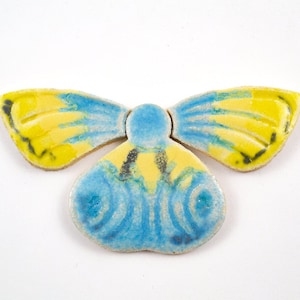 Könnte beinhalten: Eine Keramikfliese in Form eines Schmetterlings mit gelber und blauer Glasur. Der Schmetterling hat einen stilisierten Körper und Flügel mit einem Wirbelmuster.