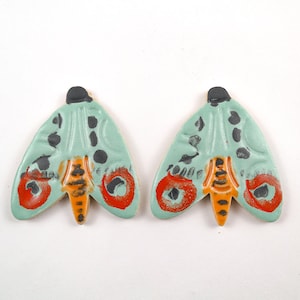 Könnte beinhalten: Ein Paar Keramik-Ohrringe in Form eines Nachtfalters mit tealblauer Basis, orangefarbenen und schwarzen Akzenten sowie roten und schwarzen Details.