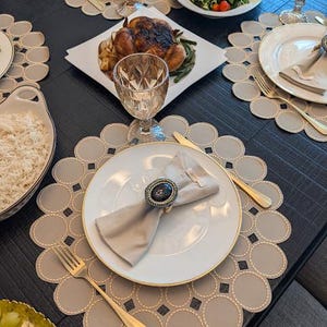 May include: A dining table set for a meal, featuring white plates with gold rims, gold cutlery, and patterned placemats. A roasted chicken, salad, and rice are visible. Crystal glasses and a napkin ring add elegance.