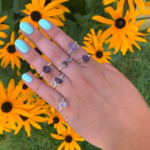 May include: Five silver rings with amethyst stones wrapped in wire. The rings are worn on a hand with light blue nail polish.