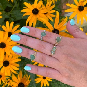 May include: A hand wearing four rings with green stones wrapped in gold wire. The rings are on the index, middle, ring, and little fingers. The hand is against a background of yellow flowers.