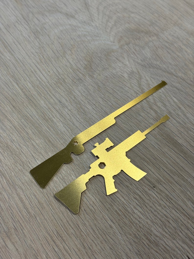 May include: Two gold metal cutouts of rifles. The top cutout is a long rifle with a scope. The bottom cutout is a shorter rifle with a scope.