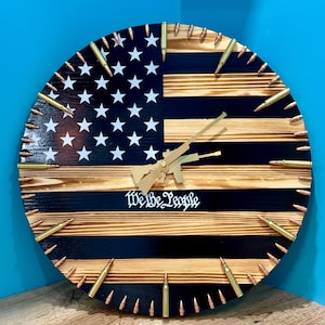 May include: A wooden clock with a black and brown American flag design. The clock is decorated with gold bullet casings around the edge. The clock face has a gold rifle design and the words "We the People" in gold lettering.