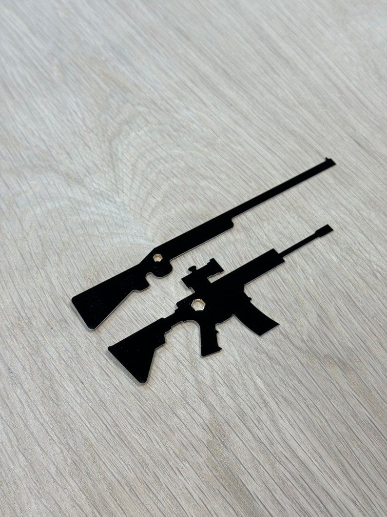 May include: Two black metal silhouette cutouts of rifles. The rifles are positioned on top of each other, with the larger rifle on top.