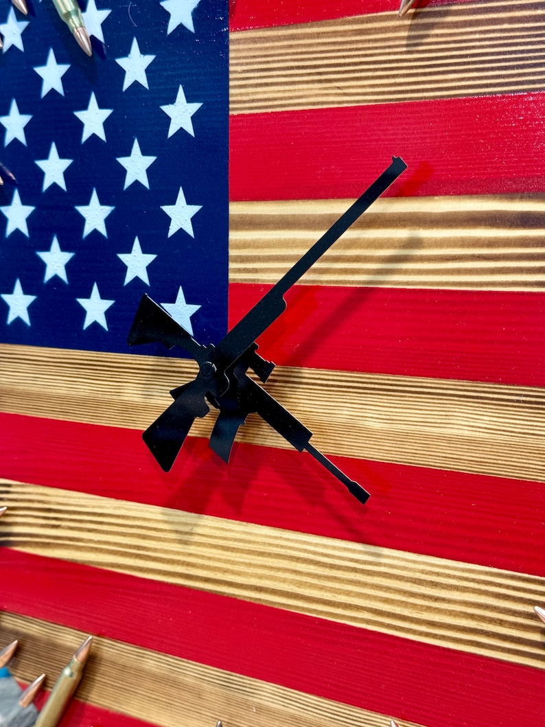 Peut inclure: Un drapeau am&eacute;ricain en bois avec un design rouge, blanc et bleu. Le drapeau est d&eacute;cor&eacute; de douilles de balles et a une silhouette noire d'un fusil comme aiguilles d'horloge.