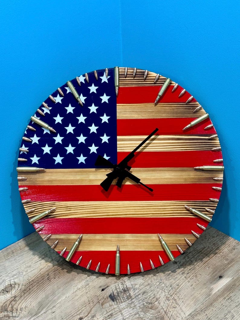Peut inclure: Une horloge ronde en bois avec un design de drapeau am&eacute;ricain rouge, blanc et bleu. Le cadran de l'horloge est entour&eacute; de douilles de balles. Les aiguilles de l'horloge sont noires et en forme de fusil.