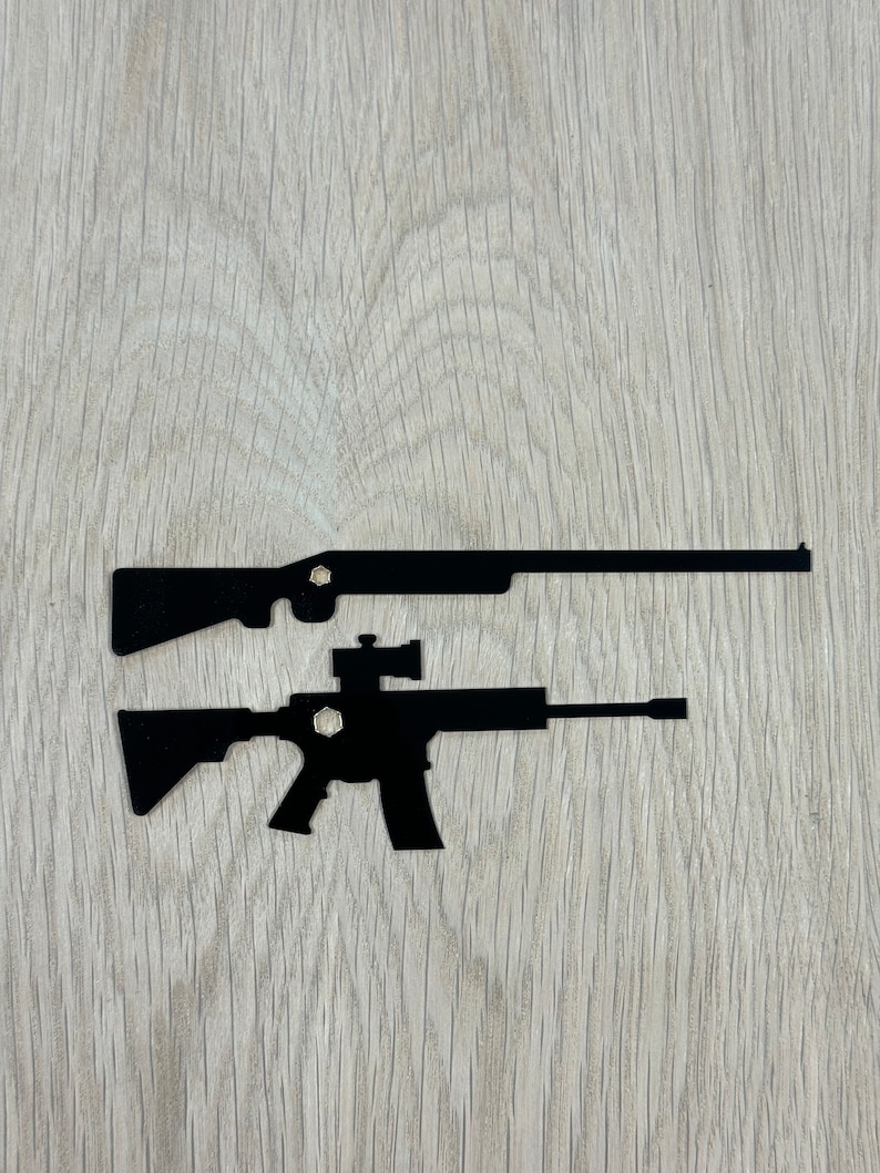 May include: Two black metal silhouettes of rifles on a light wood background. The top rifle is a shotgun and the bottom rifle is an assault rifle.