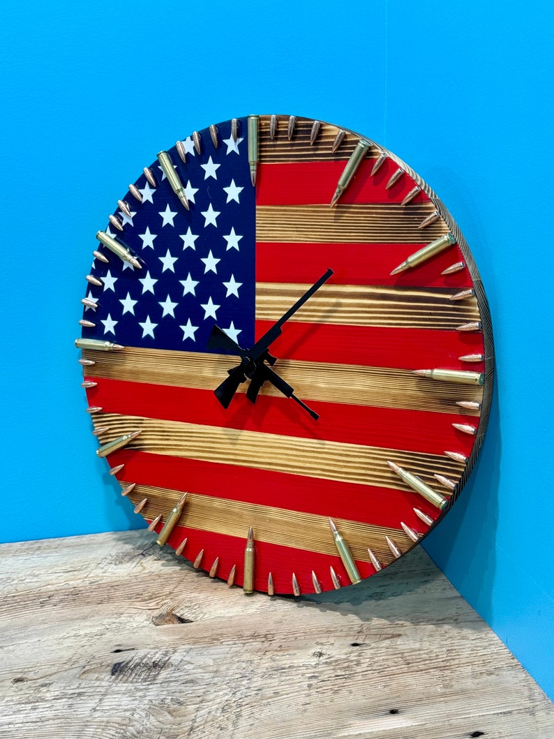 Peut inclure: Une horloge ronde en bois avec un motif de drapeau am&eacute;ricain. Le cadran de l'horloge est en bois avec des rayures rouges et blanches et des &eacute;toiles bleues. Les aiguilles de l'horloge sont noires et les chiffres sont en douilles de balles en laiton.
