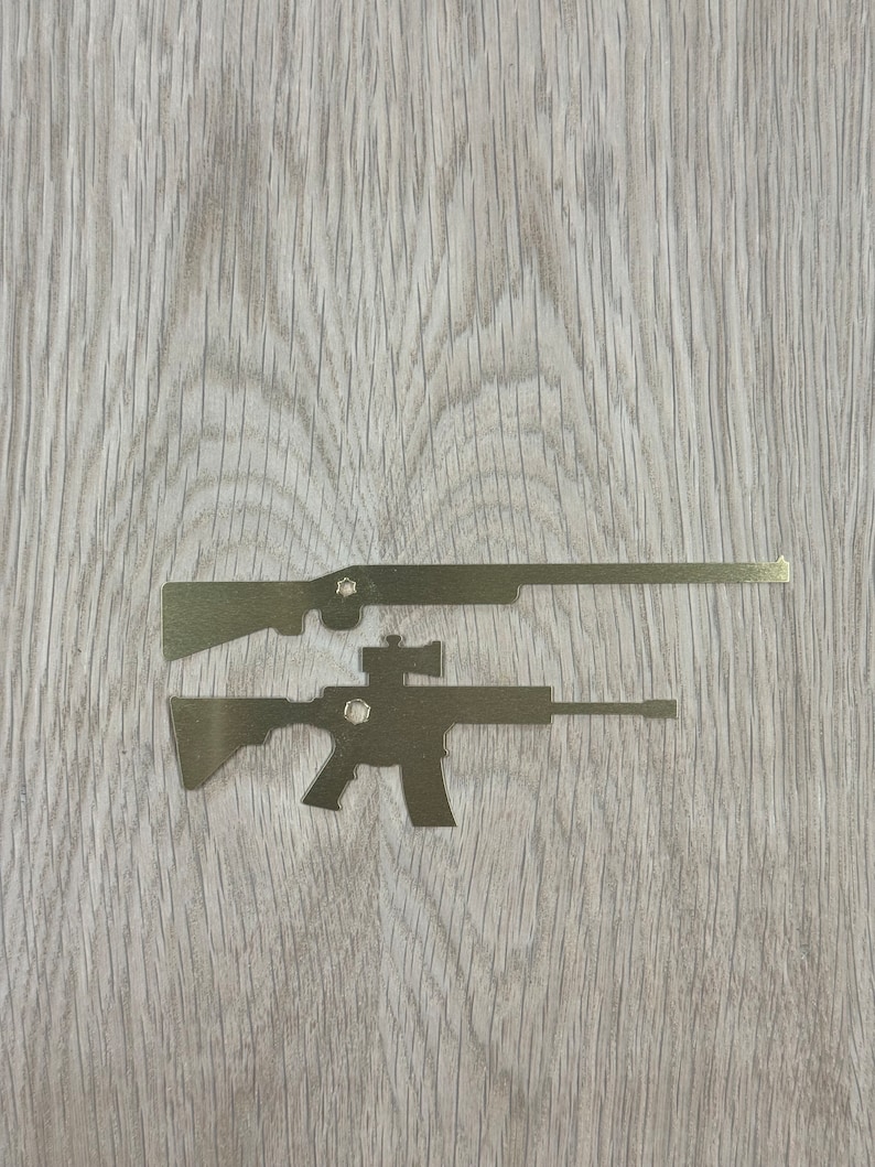 May include: Two metal silhouettes of rifles on a woodgrain background. The top rifle is a long gun with a scope. The bottom rifle is a shorter rifle with a scope.