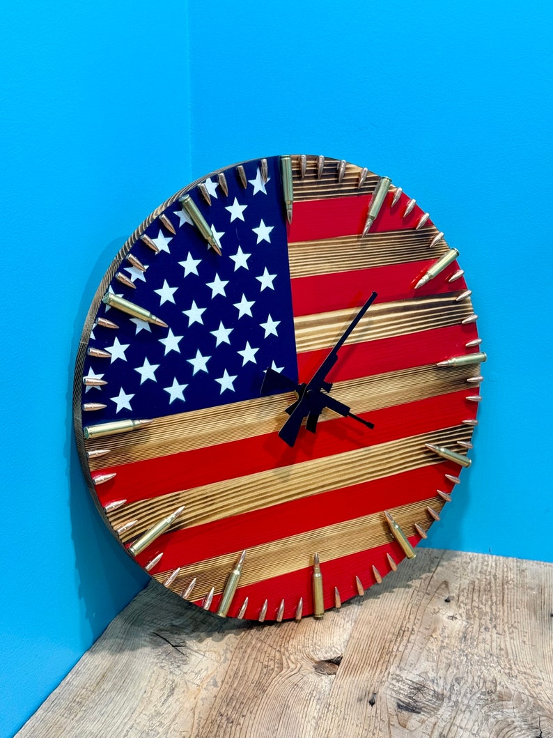 Peut inclure: Une horloge en bois avec un design de drapeau am&eacute;ricain. Le cadran de l'horloge est en bois et comporte des douilles de balles autour du bord. Les aiguilles de l'horloge sont noires et le cadran est rouge, blanc et bleu.