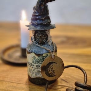 Könnte beinhalten: Eine dekorative Flasche mit einem Hexenhut-Verschluss, einem Wachssiegel und einem Anhänger mit einem Mondsichel-Design. Die Flasche hat eine Vintage-Ästhetik mit einem Etikett mit Text. Eine brennende Kerze ist im Hintergrund. Die Flasche ist etwa 15 cm hoch.