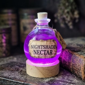 Könnte beinhalten: Eine runde Glasflasche gefüllt mit leuchtend violetter Flüssigkeit, verschlossen mit einem Korken. Ein Papieretikett trägt die Aufschrift NIGHTSHADE NECTAR in schwarzer Schrift. Die Flasche steht auf einem runden, beleuchteten Holzsockel.