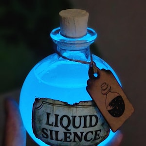 Peut inclure: Une bouteille en verre ronde remplie d'un liquide bleu lumineux, scellée par un bouchon en liège. Une étiquette en papier vieilli sur le devant porte l'inscription "LIQUID SILENCE" en noir. Une petite étiquette rectangulaire beige pend au goulot.