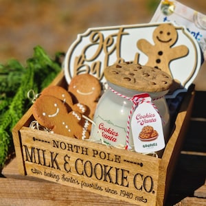 Könnte beinhalten: Eine Holzkiste mit der Aufschrift North Pole Milk & Cookie Co. enthält eine Kerze mit einem keksförmigen Deckel, Lebkuchenmänner und ein dekoratives Schild mit einem Lebkuchenmann und dem Text get baked. Ein Etikett zeigt Cookies for Santa.