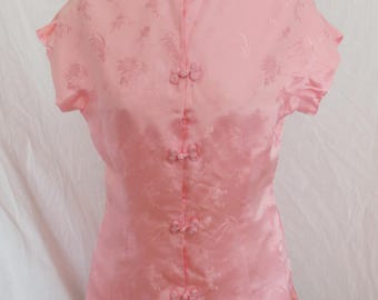 May include: A light pink, short-sleeved blouse with a mandarin collar and decorative frog closures. The blouse features a subtle floral pattern and is displayed on a mannequin. The style is reminiscent of traditional Chinese clothing.
