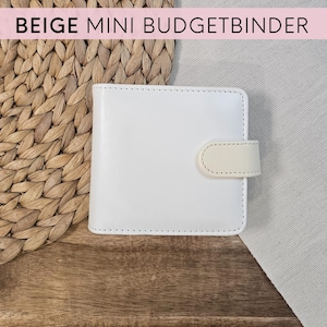 Könnte beinhalten: Ein weißer Mini-Budget-Ordner mit einem cremefarbenen Verschluss. Der Ordner liegt auf einer Holzoberfläche neben einer gewebten Matte und einem grauen Stoff. Der Text "BEIGE MINI BUDGETBINDER" steht oben.