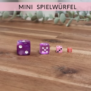Può includere: Quattro dadi in miniatura di diversi colori: viola, trasparente, rosa e rosso. I dadi sono su una superficie di legno. Il testo "MINI SPIELWÜRFEL" è visibile nella parte superiore dell'immagine.