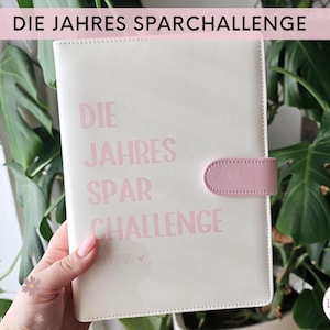 Könnte beinhalten: Ein weißes, ledergebundenes Journal mit einer rosa Lasche und dem Text "DIE JAHRES SPAR CHALLENGE" in rosa Schrift.