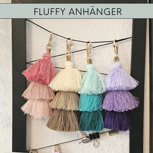 May include: Five multi-colored fluffy tassel keychains with gold metal rings. The tassels are layered in three tiers and are pink, cream, light blue, lavender, and purple.