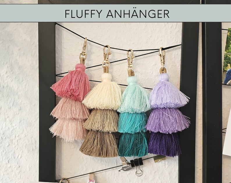 May include: Five multi-colored fluffy tassel keychains with gold metal rings. The tassels are layered in three tiers and are pink, cream, light blue, lavender, and purple.