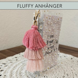 May include: A pink, three-tiered tassel keychain with a gold ring attached to a clear plastic notebook with a white daisy pattern and the text "Mein Bucha - Meine Geschichten".