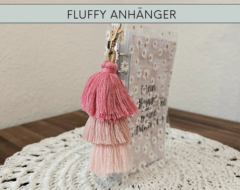 May include: A pink, three-tiered tassel keychain with a gold ring attached to a clear plastic notebook with a white daisy pattern and the text "Mein Bucha - Meine Geschichten".