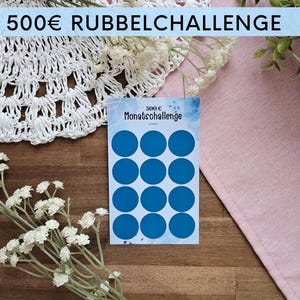 Może przedstawiać: Karta z dwunastoma niebieskimi okręgami, oznaczona jako "300€ Monatsschallenge", jest wyświetlana na drewnianej powierzchni. Tekst "500€ RUBBELCHALLENGE" znajduje się u góry. Karta jest otoczona białymi kwiatami, białą koronkową serwetką i różową tkaniną.