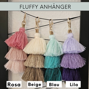 May include: Four fluffy tassel keychains in pink, beige, blue, and purple. The keychains are layered with three tassels each, and they are hanging from gold clasps. The text "FLUFFY ANH&Auml;NGER" is at the top of the image, and the colors are labeled "Rosa", "Beige", "Blau", and "Lila" at the bottom.