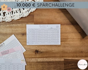 Könnte beinhalten: Spar-Challenge-Karten mit dem Text "10.000 € Sparchallenge". Die Karten zeigen Raster mit Zahlen zur Verfolgung von Sparzielen. Sie liegen auf einer Holzoberfläche, mit einer dekorativen Häkelarbeit und einer strukturierten grauen Oberfläche.