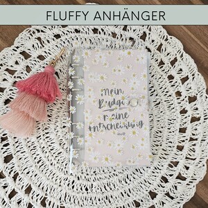 May include: A pink and white fluffy tassel keychain attached to a clear plastic planner with a white daisy pattern and the text "Mein Budget ist keine Entschuldigung".