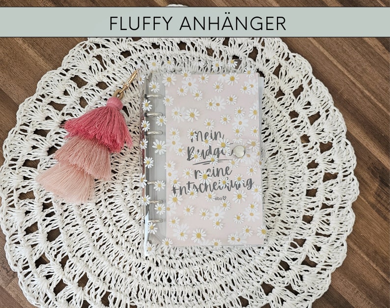 May include: A pink and white fluffy tassel keychain attached to a clear plastic planner with a white daisy pattern and the text "Mein Budget ist keine Entschuldigung".