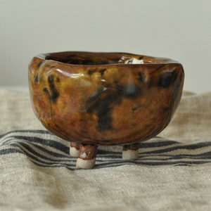 May include: A small, brown ceramic bowl with a mottled glaze. The bowl has three short legs and is sitting on a striped fabric.